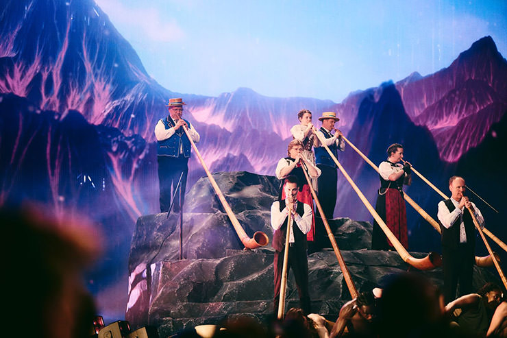 Musicians from Lochus Alphorn Quartett performing Eurovision medley at the opening of the 69th Eurovision Song Contest in Basel, with magenta-colored mountains displayed on a LED wall in the background; photo by Alma Bengtsson / EBU.
