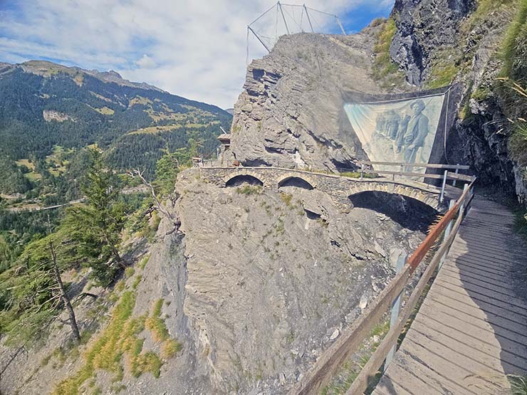 Wooden path leading to the stone bridge, followed by a metal suspended bridge, along the Bisse du Torrent Neuf, a hiking trail in Saviese, Switzerland, there is a large vintage poster showing an image of the locals in the 1930s balancing on the wooden planks above the abyss; photo by Ivan Kralj.