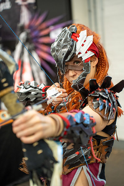 Swiss cosplayer Miri, known as Secret Geek, pointing a bow and an arrow towards the camera, while cosplaying Aloy from the video game series "Horizon"; copyright Fantasy Basel, the Swiss Comic Con.