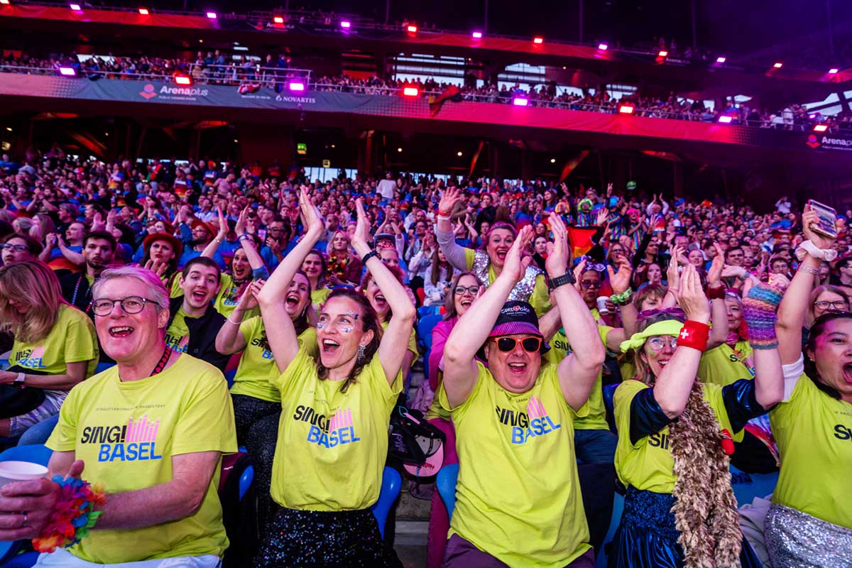 Audience in "Sing Basel" T-shirts clapping and cheering at St. Jakob-Park stadium in Basel during Arena Plus Eurovision pre-show; photo by Mood Studios AG.