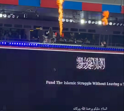 The view of a brief on-screen glitch flashing "Fund the Islamic Struggle Without Leaving a Trace" behind Baby Lasagna drummer at St. Jakob-Park stadium in Basel, Switzerland, during Arena Plus Eurovision pre-party; screenshot by Pipeaway.