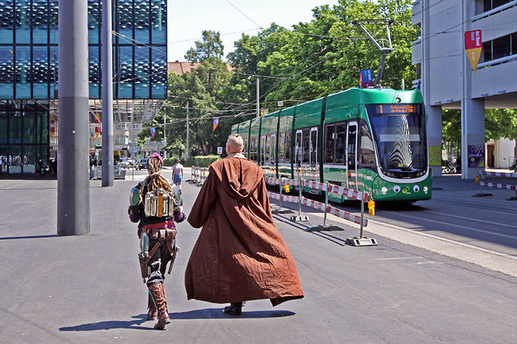 Shot from the back side, cosplayers recognizably dressed as Boba Fett and Obi-Wan Kenobi from "Star Wars" walking over Messeplatz, with a tram number 1 in the background, heading to Fantasy Basel, the Swiss Comic Con; photo by Ivan Kralj.