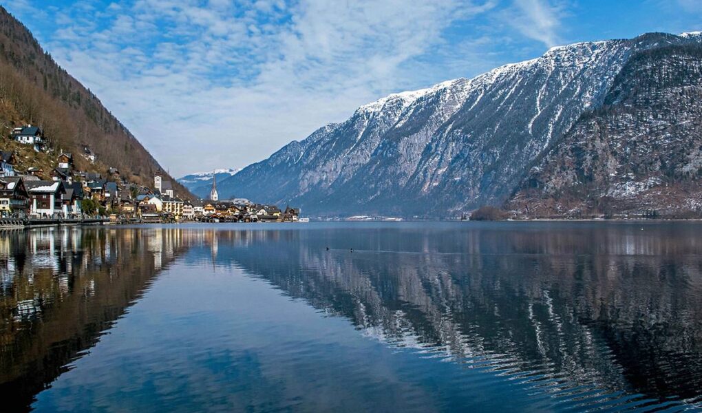 Reflective surface of Hallstätter See, lake by the picturesque Hallstatt town, with snow-peaked mountain in the background; photo by Ljubomir Žarković / Unsplash.