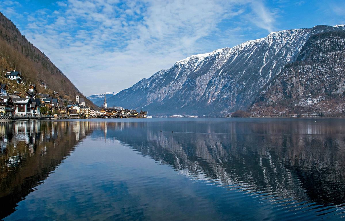 Reflective surface of Hallstätter See, lake by the picturesque Hallstatt town, with snow-peaked mountain in the background; photo by Ljubomir Žarković / Unsplash.