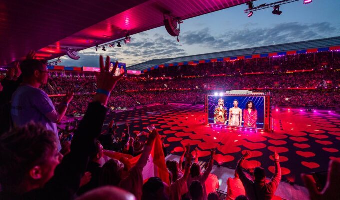 Eurovision hosts appearing on a gigantic cube screen at St. Jakob-Park, with delighted Arena Plus audiences; photo by Mood Studios AG, Kanton Basel-Stadt.