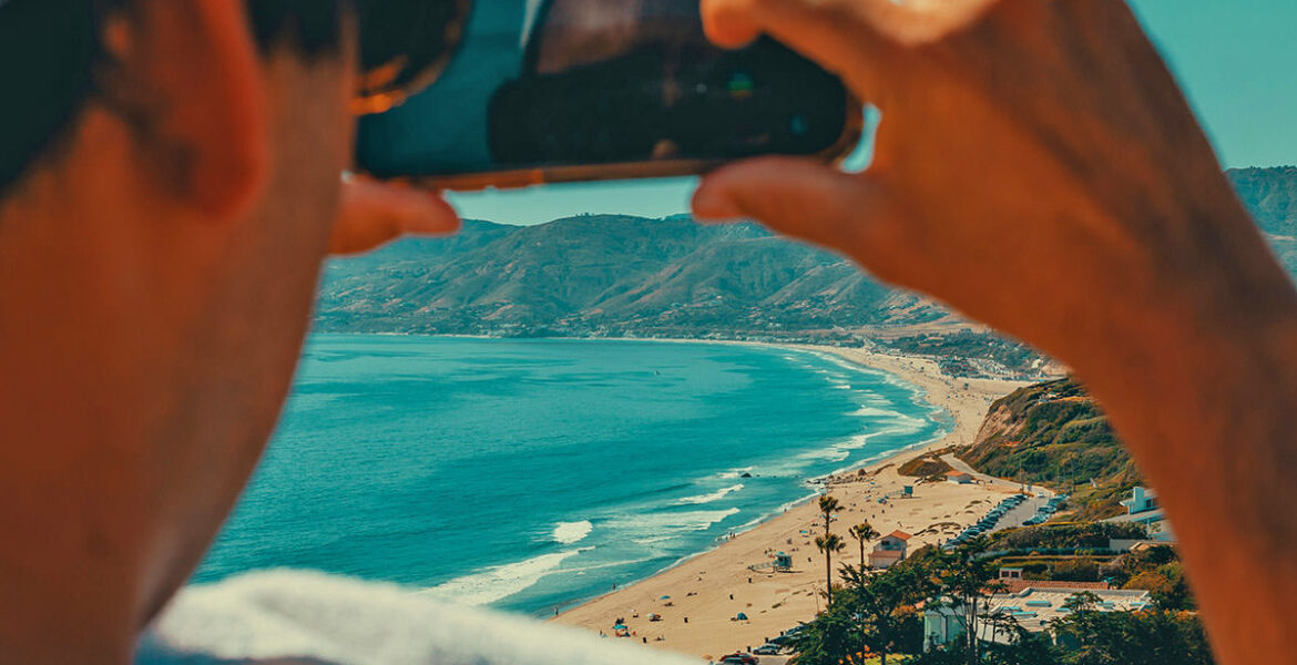 A man taking a travel video of a beach in Malibu with his phone; photo by Tom Briskey, Unsplash.