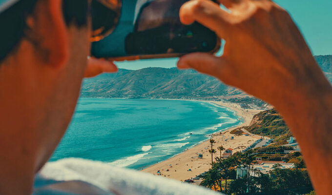 A man taking a travel video of a beach in Malibu with his phone; photo by Tom Briskey, Unsplash.