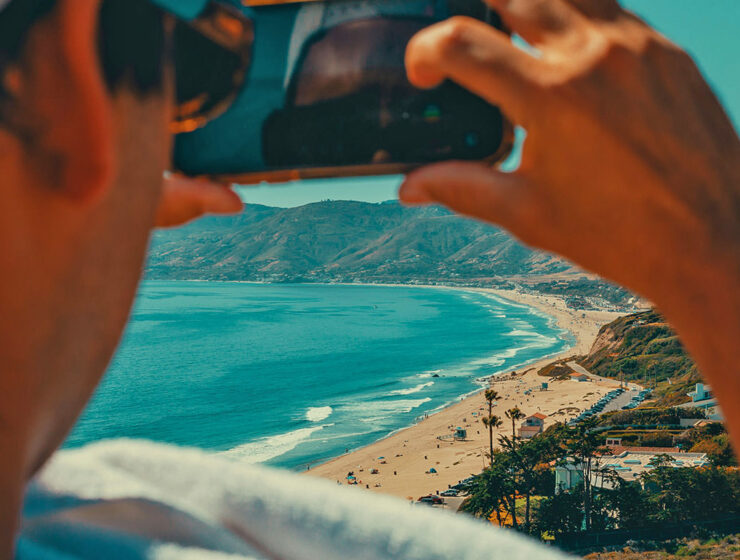 A man taking a travel video of a beach in Malibu with his phone; photo by Tom Briskey, Unsplash.