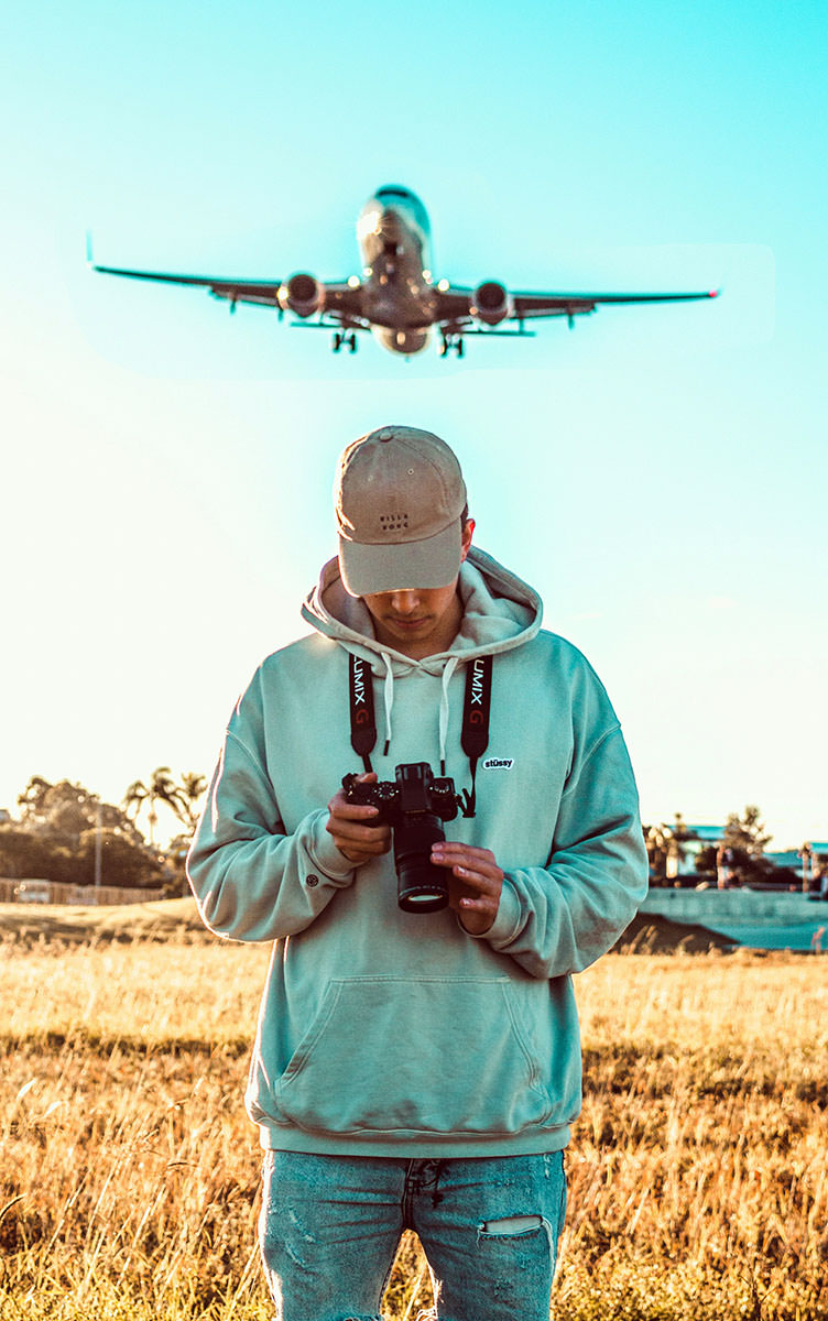 A plane flying over a man with a camera looking downwards while standing on a field; photo by Justin Clark, Unsplash.