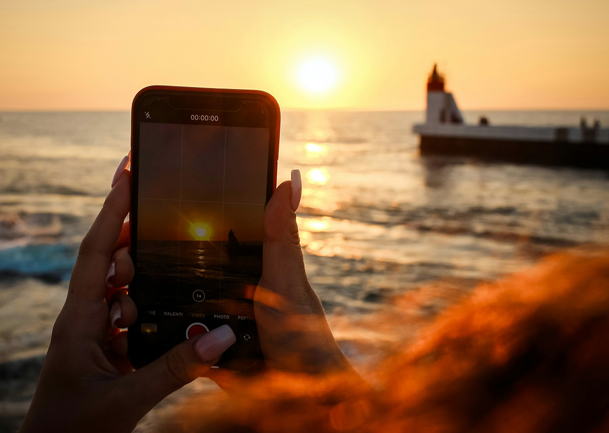 Long-nailed hands holding a smartphone shooting a video of a sunset over the sea horizon; photo by Jametlene Reskp, Unsplash.