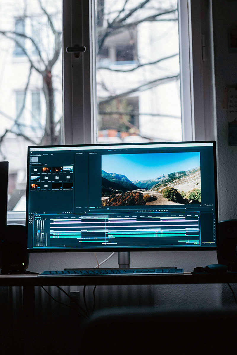 Flat screen computer monitor showing a travel video editing software, located in a room, with the window looking at urban environment behind; photo by Danny Feng, Unsplash.