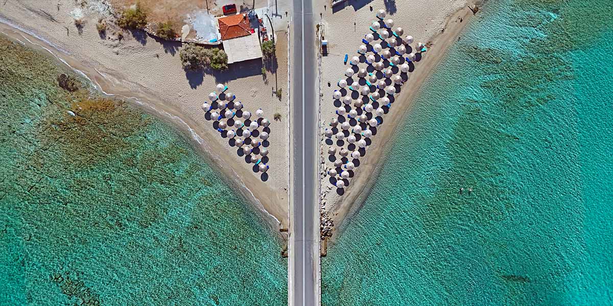 Aerial view of sandy beaches with parasols and turquoise water wrapping the road bridge in Diakofti, Kythira Island, Greece; photo by Ivan Kralj.