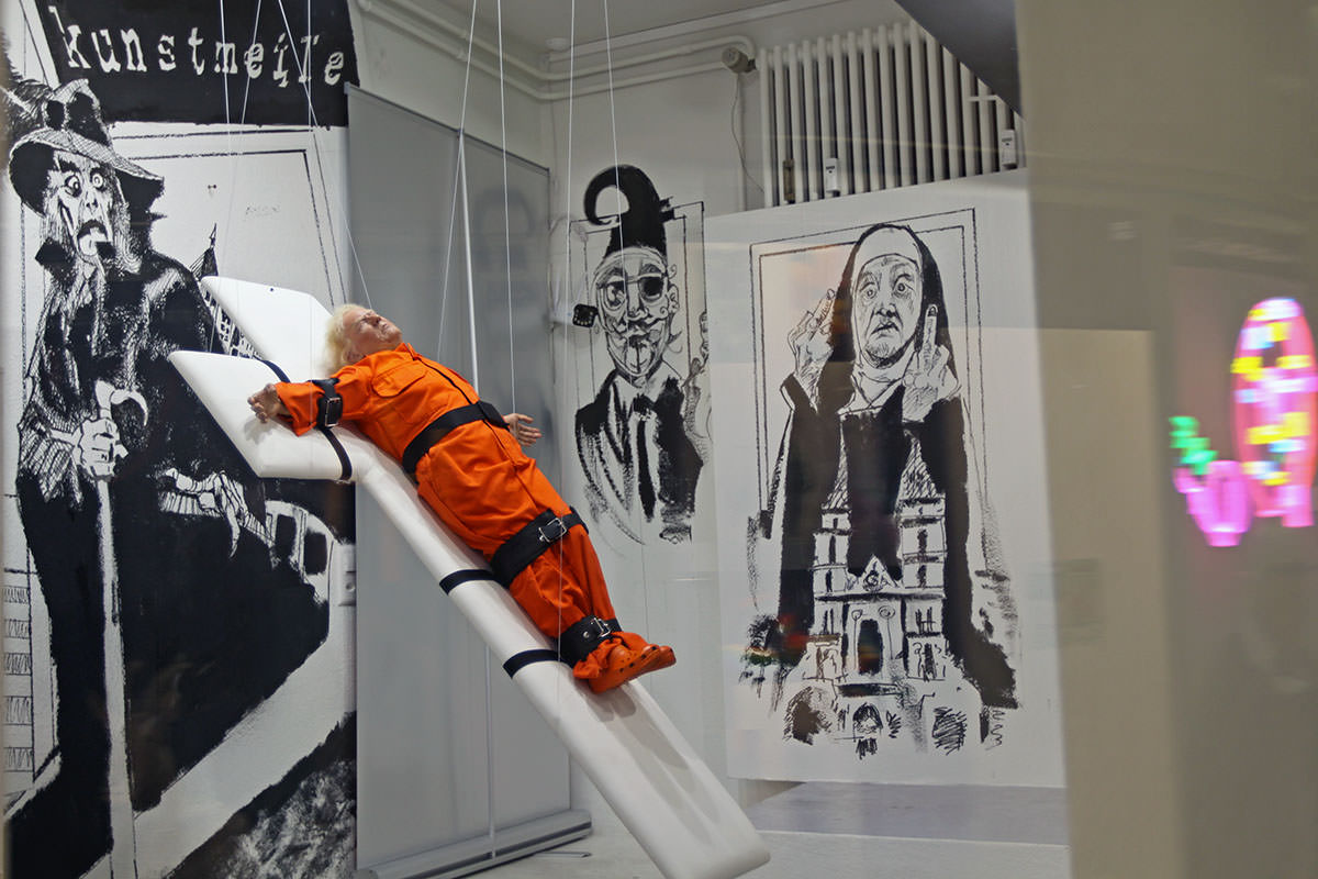 A figure of Donald Trump crucified in the exhibition window of Kunstmeile Basel, with black-and-white finger-flipping nun on the mural in the background - Trump strapped to a white cross in orange prison jumpsuit is an artwork "Saint or Sinner?" by Mason Storm; photo by Ivan Kralj. 