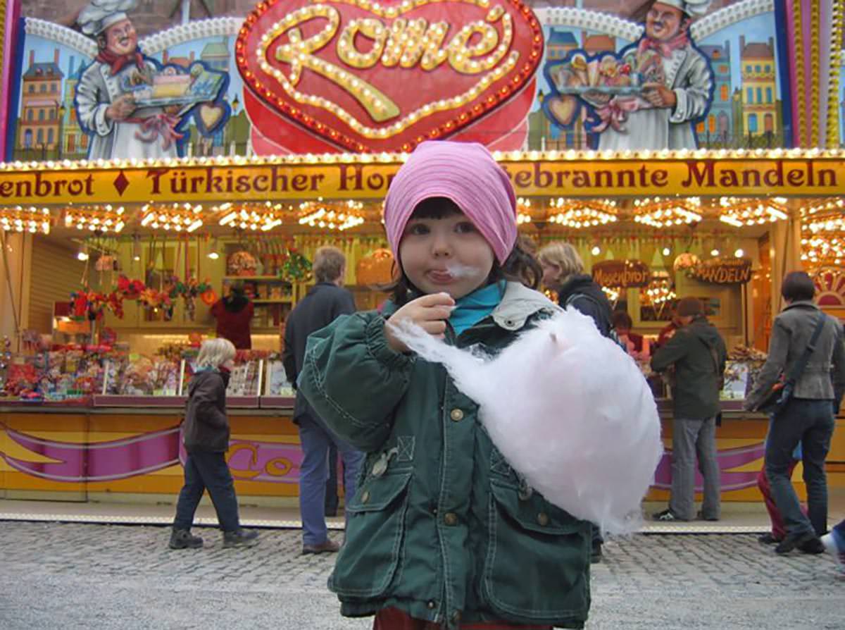 A little girl eating a pink cotton candy while standing in front of "Rome" confiserie at Basler Herbstmesse, Basel Autumn Fair in Switzerland; photo by Georg Holderier, Polapix.