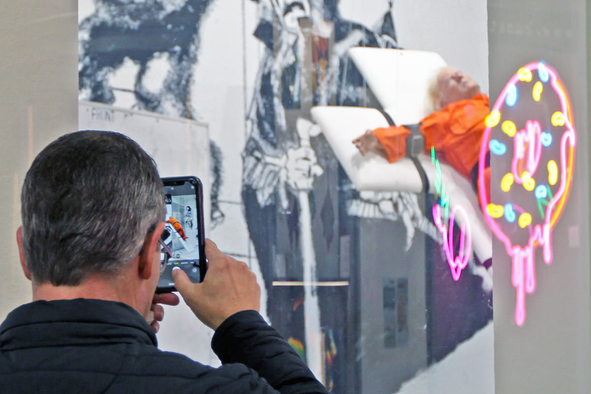 A close-up of a man photographing Mason Storm's artwork "Saint or Sinner?" with his phone, the installation showing a miniature Donald Trump strapped to a cross in an orange prison jumpsuit, exhibited at Kunstmeile Basel, Switzerland; photo by Ivan Kralj.