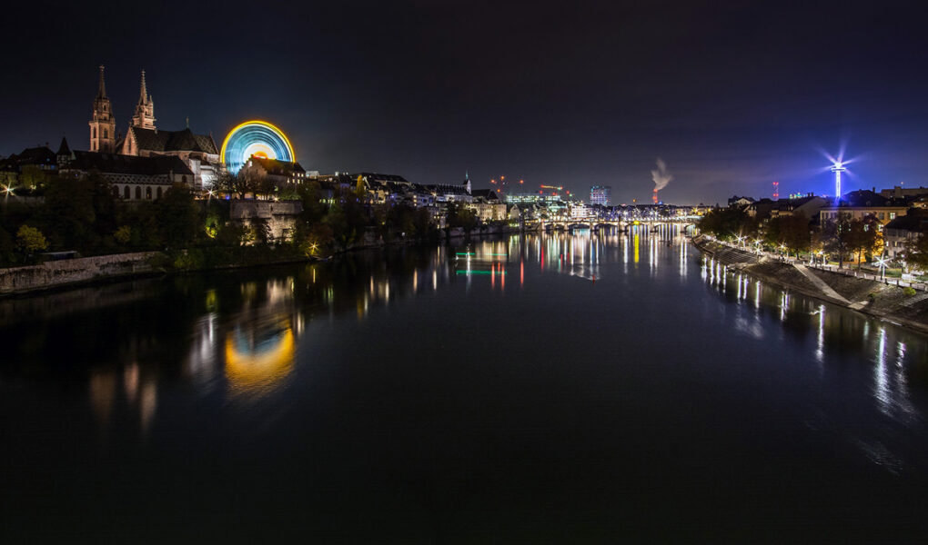 The night panorama of Rhine and Basel during Basler Herbstmesse, Basel AutumnFair, with neon-lit Ferris wheel dominating the scene at Münsterplatz; photo by Michael Kuhn.