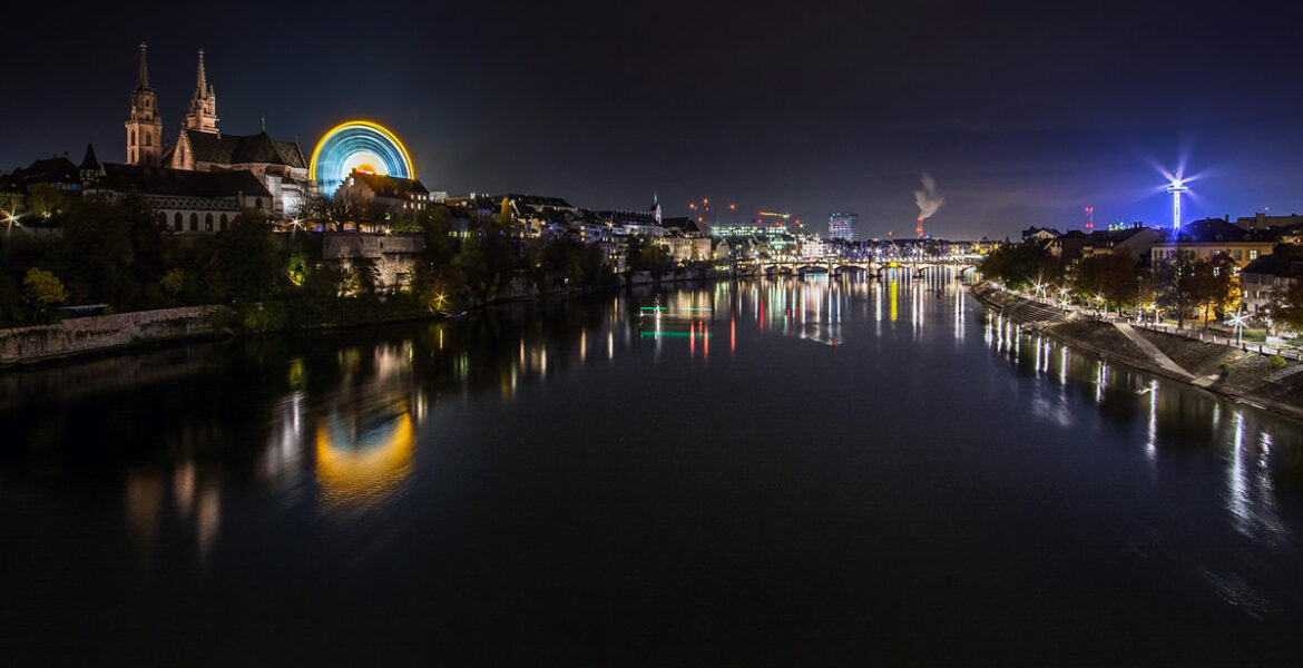The night panorama of Rhine and Basel during Basler Herbstmesse, Basel AutumnFair, with neon-lit Ferris wheel dominating the scene at Münsterplatz; photo by Michael Kuhn.