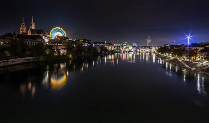 The night panorama of Rhine and Basel during Basler Herbstmesse, Basel AutumnFair, with neon-lit Ferris wheel dominating the scene at Münsterplatz; photo by Michael Kuhn.