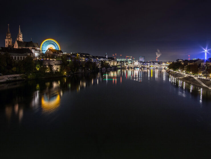 The night panorama of Rhine and Basel during Basler Herbstmesse, Basel AutumnFair, with neon-lit Ferris wheel dominating the scene at Münsterplatz; photo by Michael Kuhn.