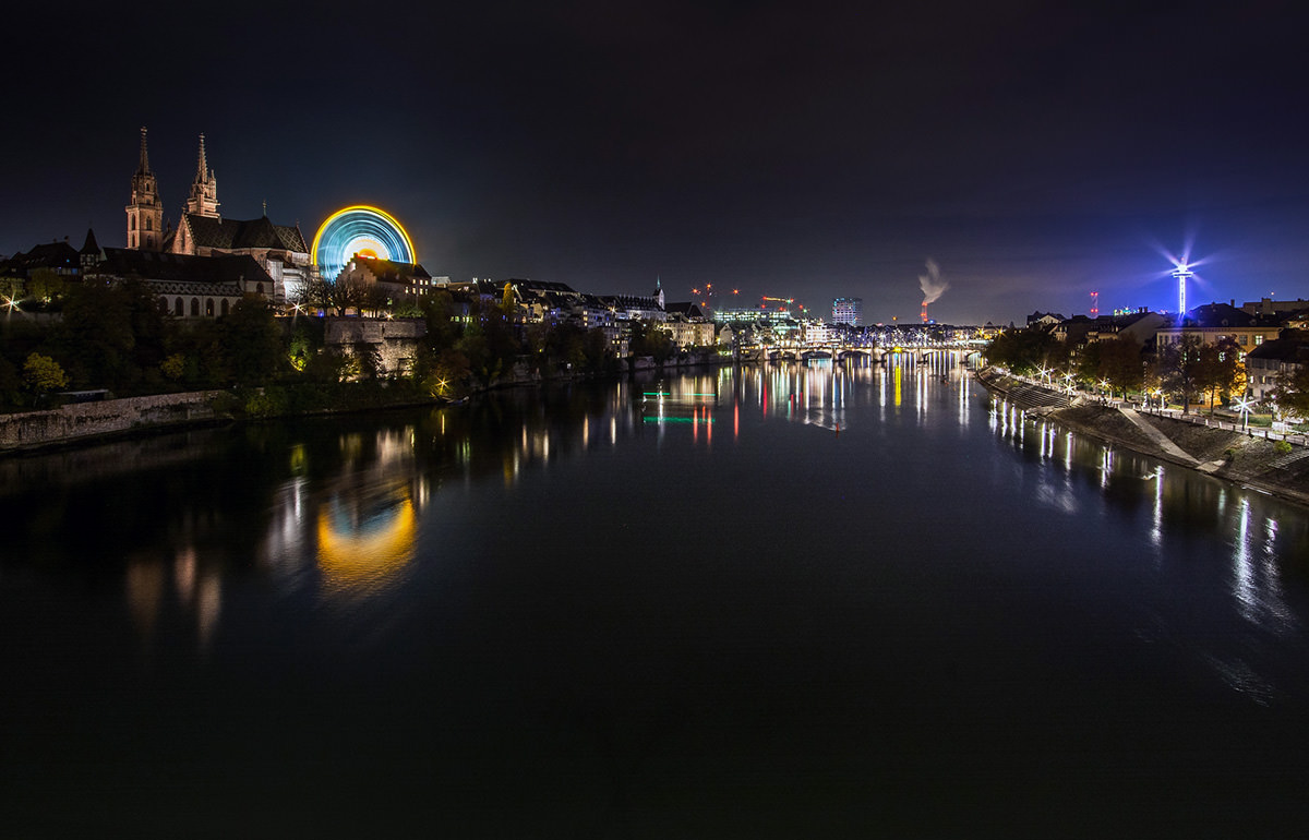 The night panorama of Rhine and Basel during Basler Herbstmesse, Basel AutumnFair, with neon-lit Ferris wheel dominating the scene at Münsterplatz; photo by Michael Kuhn.