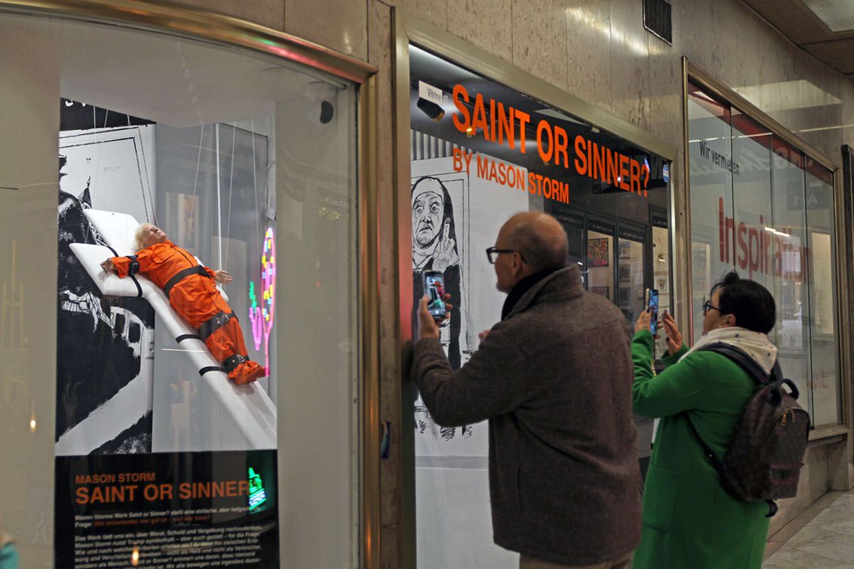 Visitors of Kunstmeile, art corridor in central Basel, photographing Mason Storm's exhibit "Saint or Sinner?", showing US President Donald Turmp dressed in an orange prison jumpsuit and strapped to a white padded cross; photo by Ivan Kralj.