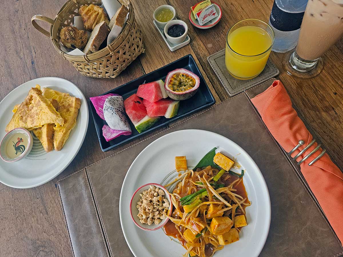 Breakfast served at The Kitchen, an on-site restaurant at Anda Mani Khao Lak Beachfront Villas in Thailand, featuring pad Thai, French toast, bread basket, fruits, and juices; photo by Ivan Kralj.