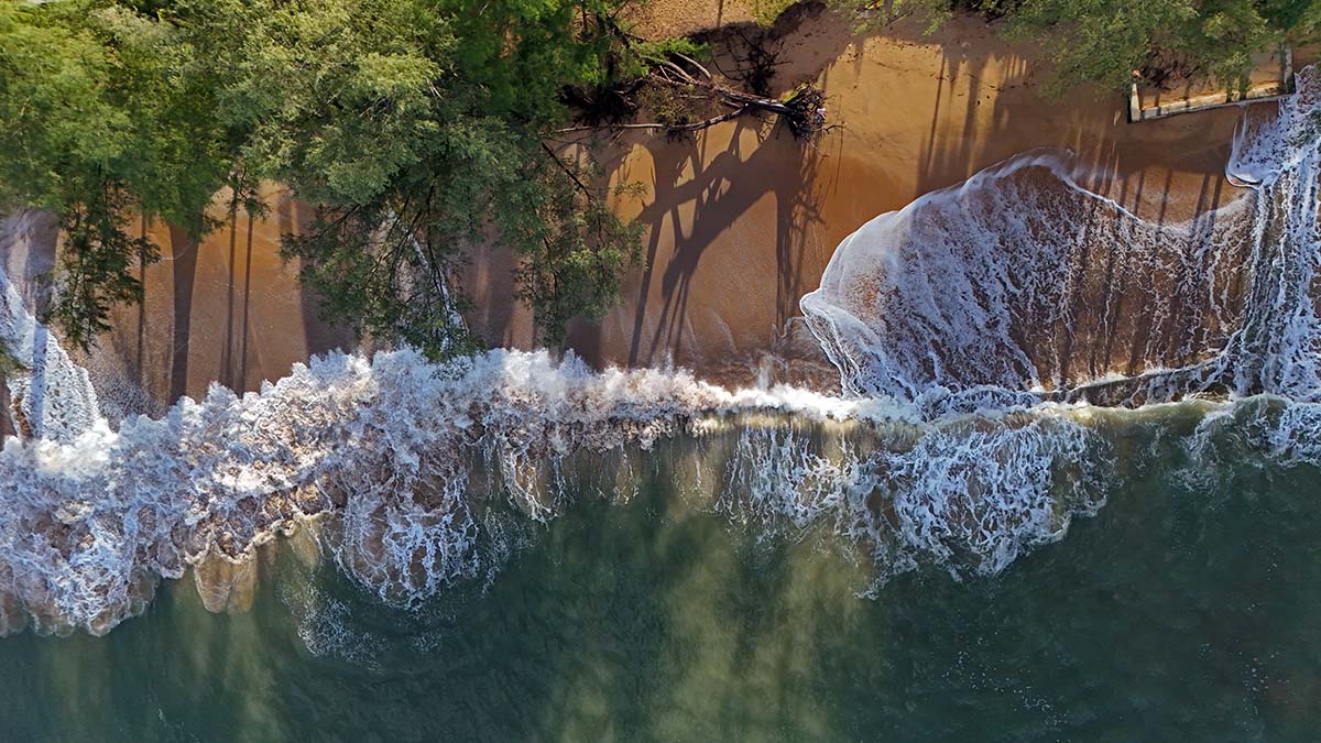 The top view of Lam Kaen Beach, with the morning sun casting long shadows of trees over Andaman Sea waves rolling the sands; drone photo by Ivan Kralj.
