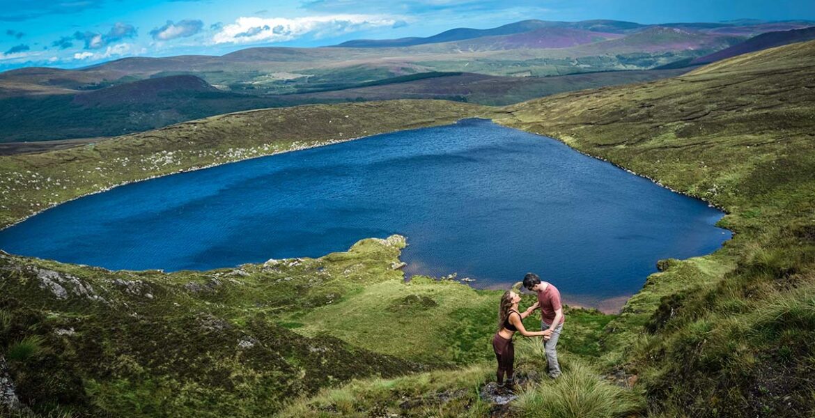 A romantic couple embracing with Lough Ouler, Ireland's heart-shaped lake in the background; photo by Dannii Coughlan, Unsplash.