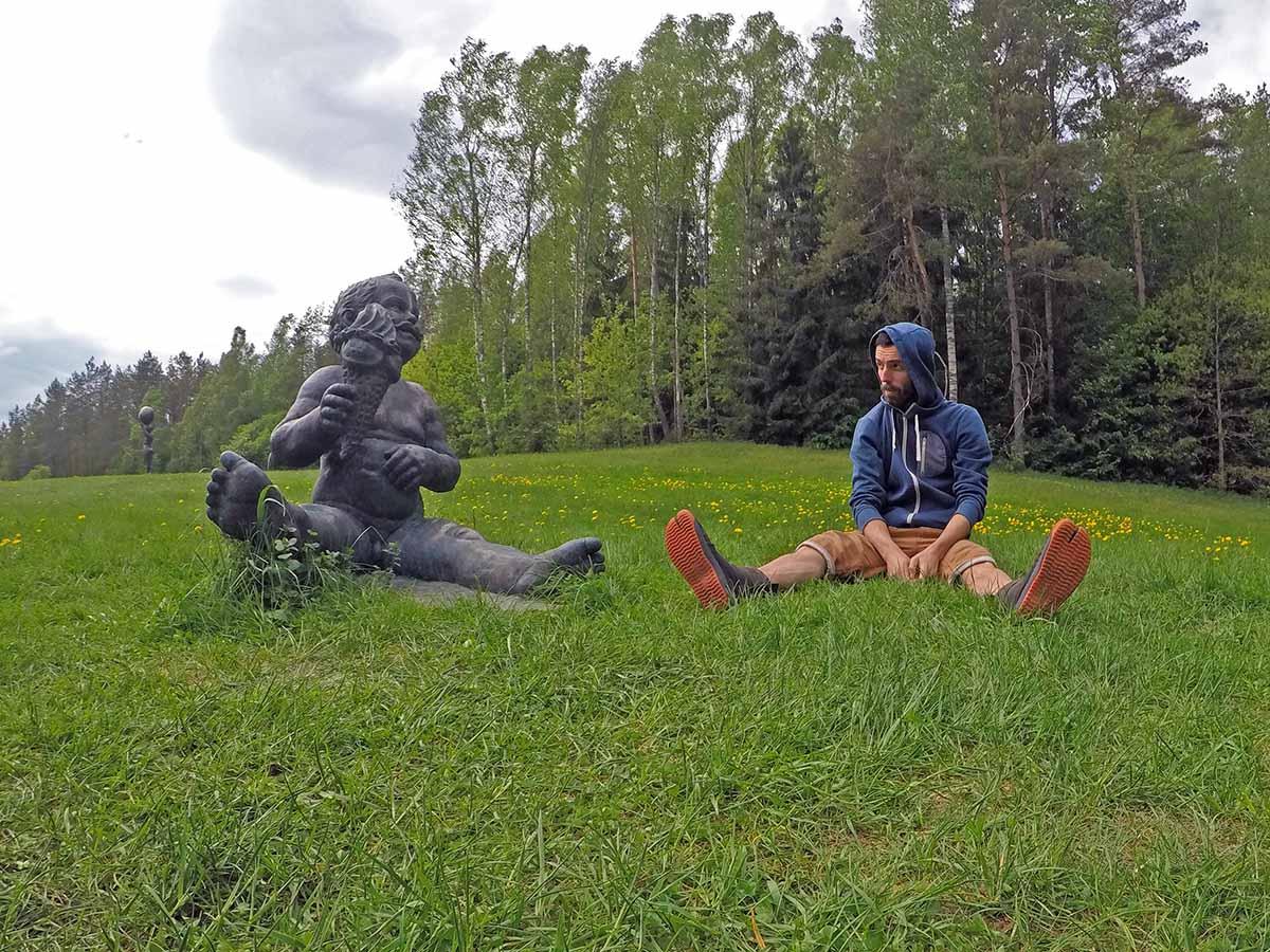 Travel blogger Ivan Kralj sitting next to a bronze boy with mustache and ice-cream ("Su Ūsais" or "With Moustaches"), an artwork by Evaldas Pauza, displayed on the meadow of Europos Parkas, the open-air museum at the geographical center of Europe, near Vilnius, Lithuania; photo by Ivan Kralj.