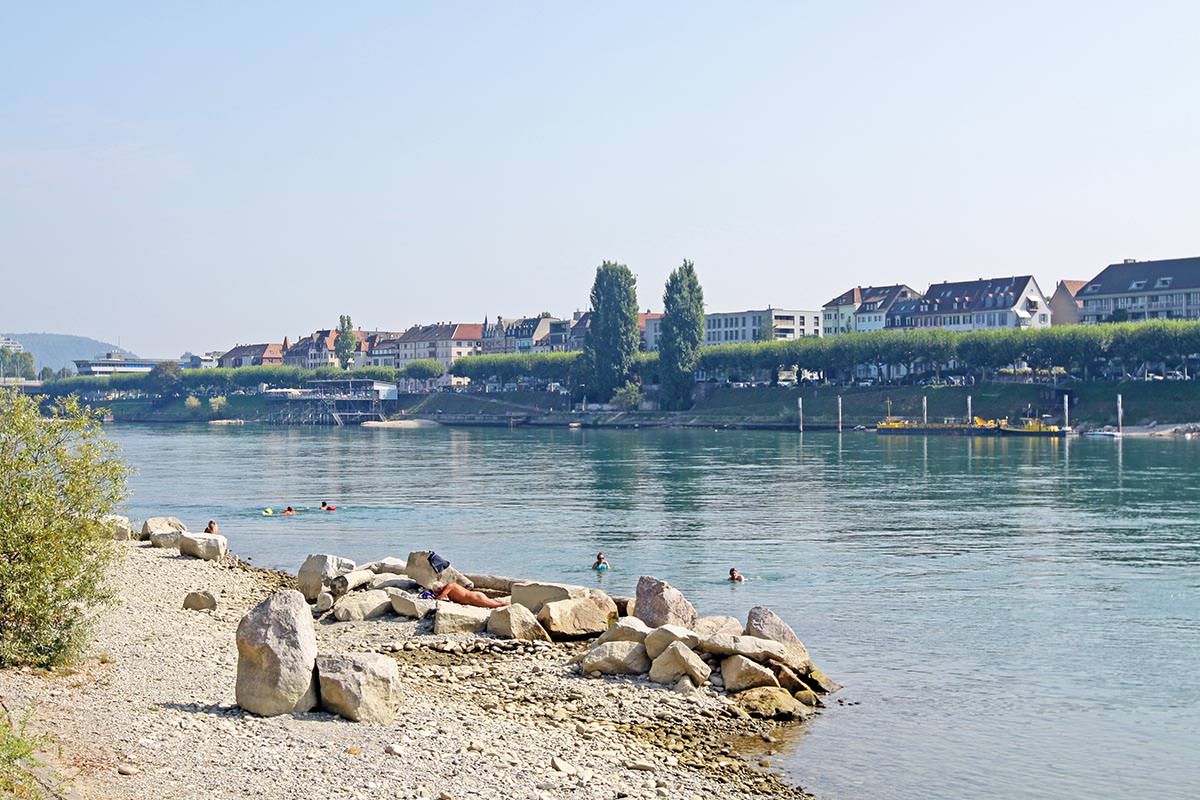 A nudist man lying on Basel's naturist beach while people with Wickelfisch bags drift by in the Rhine, Basel, Switzerland; photo by Ivan Kralj.