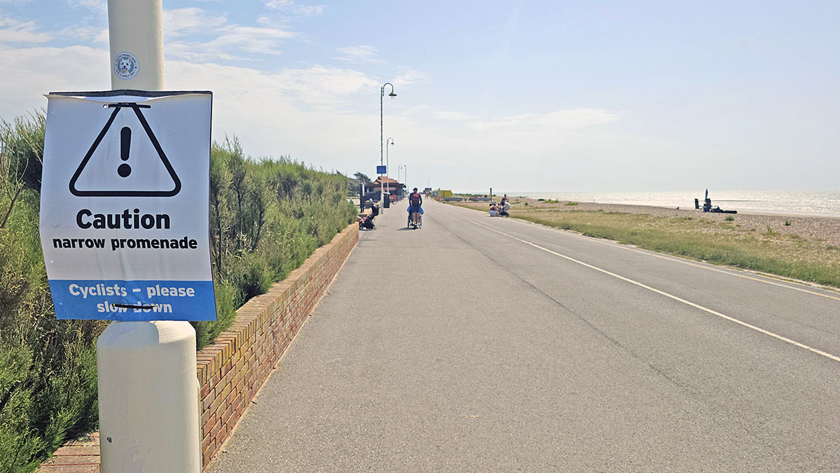 The sign on the lamppost warning "Caution, narrow promenade. Cyclists, please slow down", on a quite broad Littlehampton Beach promenade, West Sussex, England, UK; photo by Ivan Kralj.