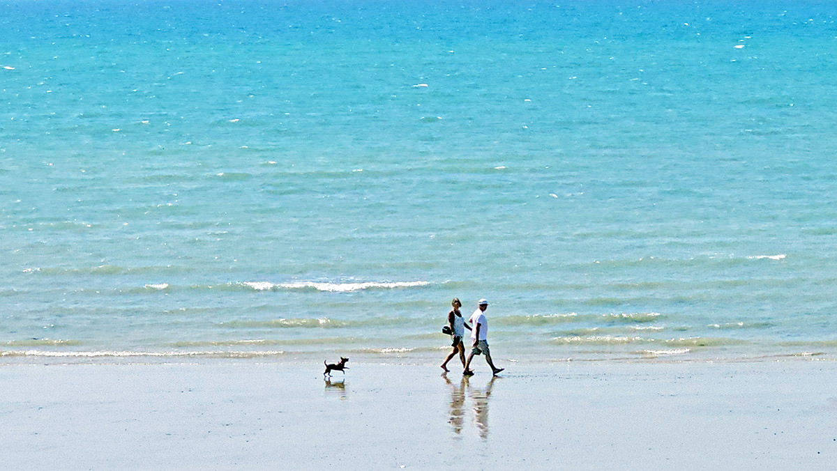 A couple walking over the sandy Littlehampton Beach, with their dog running behind them, West Sussex, England, UK; photo by Ivan Kralj.