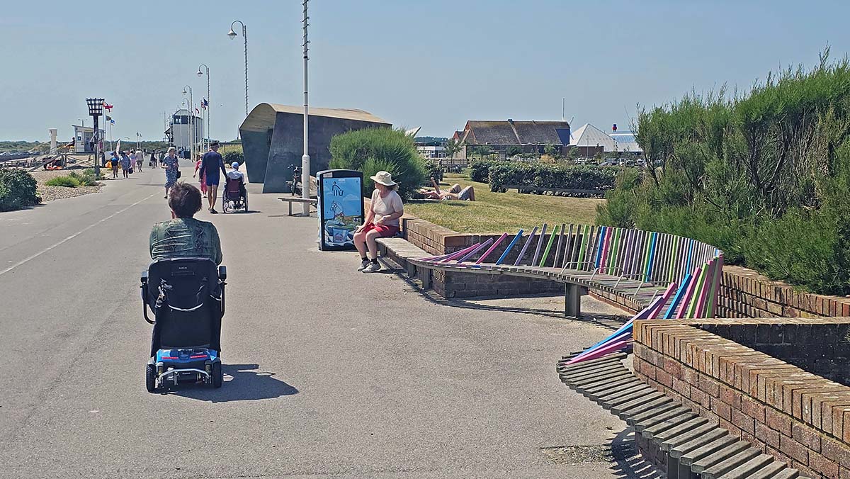 Woman sitting on the UK's longest bench while people pass by on the Littlehampton Beach promenade, with a person in the wheelchair in the foreground; photo by Ivan Kralj.